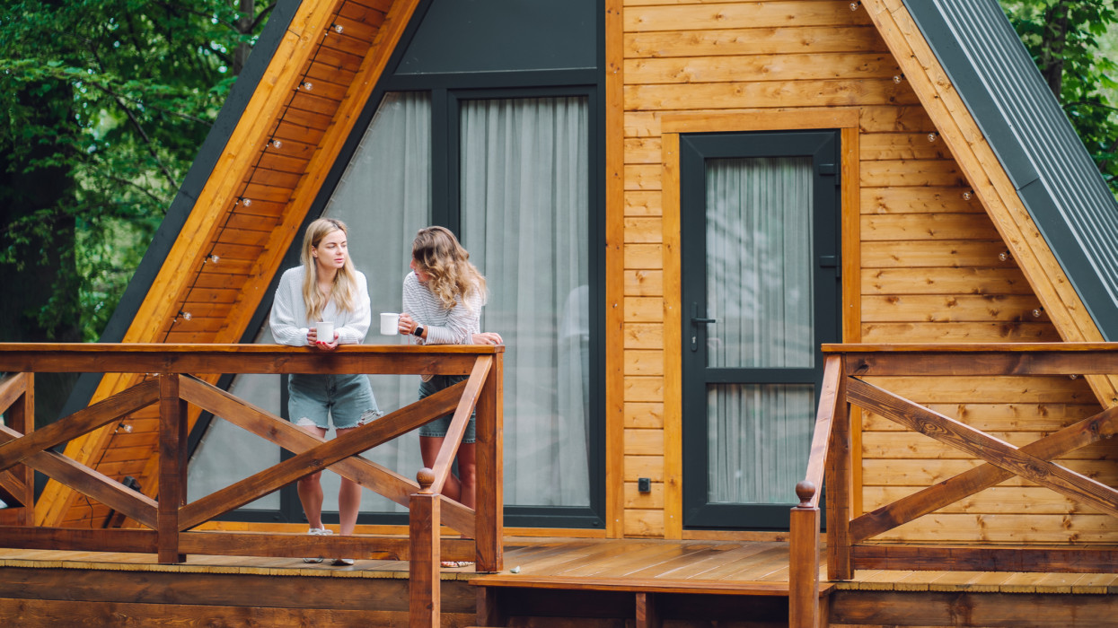 Two women drinking tea and having conversation on a wooden house balcony. Glamping getaway and time with family in tranquil atmosphere
