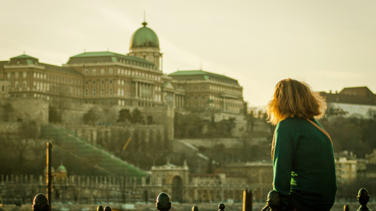 Lonely blonde girl sitting looking at the Royal Palace of Buda during sunset in Budapest, Hungary.