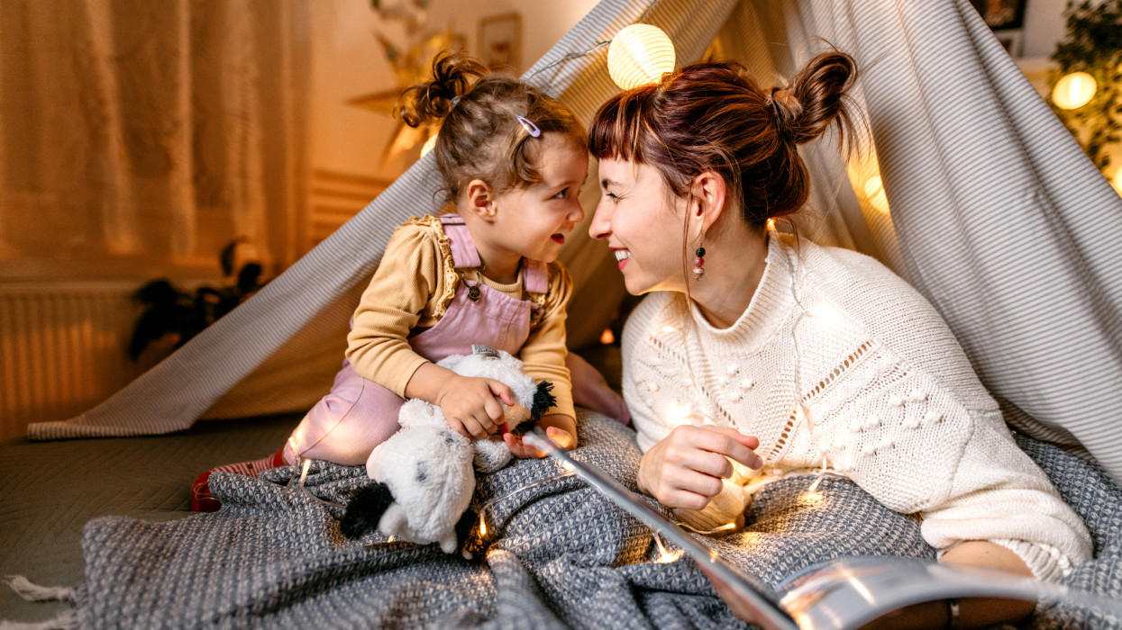 Mother and daughter are having fun reading a book under the illuminated tent in the bedroom.