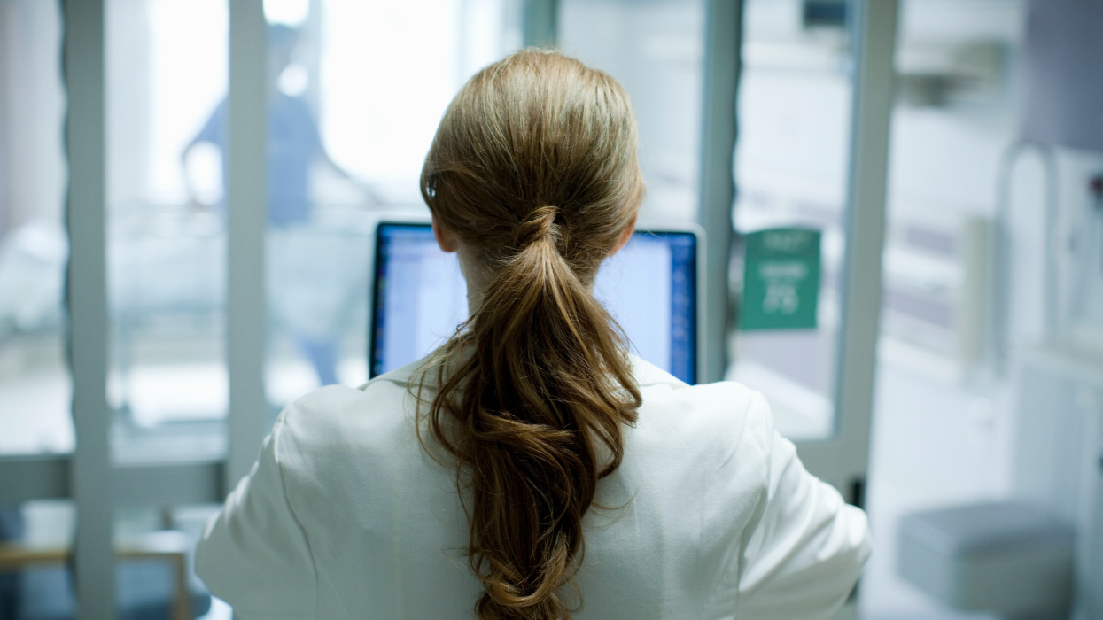 Woman doctor with long red hair using laptop in hospital, photographed from behind