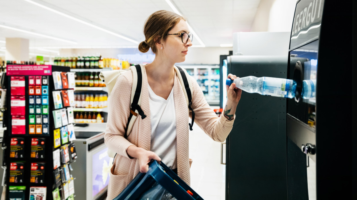 A young woman holding a crate and putting her empty plastic bottles in the recycling at her local supermarket.