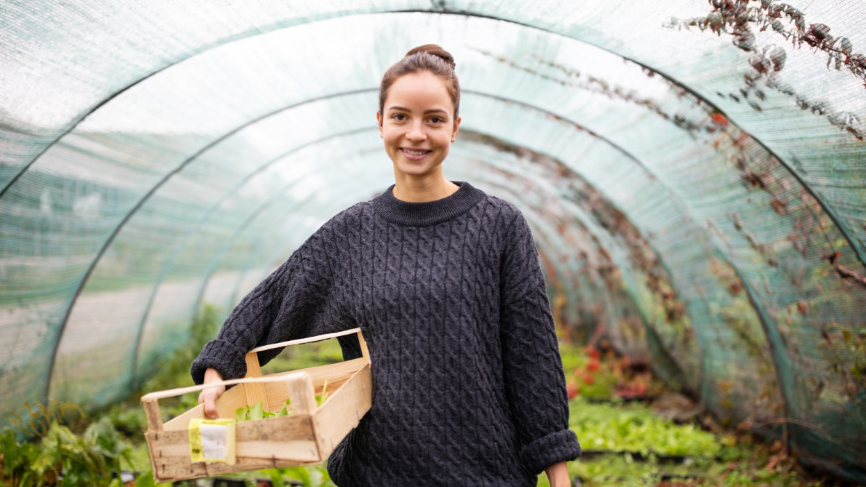 Portrait of happy young woman with wooden box standing in greenhouse. Female worker working in garden center, looking at camera and smiling.