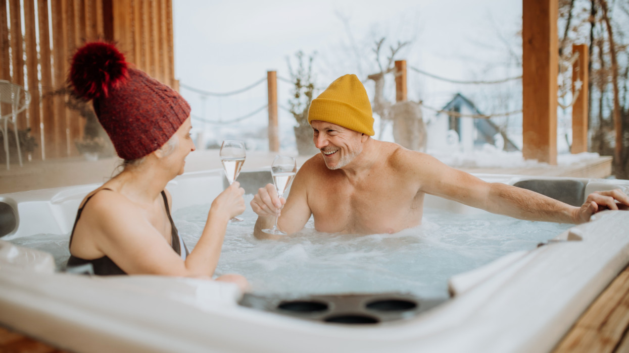 Senior couple in kintted cap enjoying together outdoor bathtub and clinking glasses at their terrace during a cold winter day.