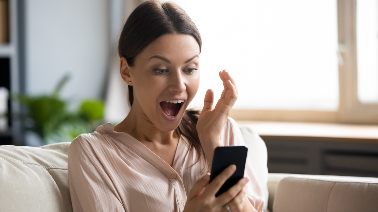 Amazed young woman sit on couch at home feel shocked reading unexpected good news on cellphone gadget, stunned millennial girl surprised by online lottery win on smartphone, luck concept