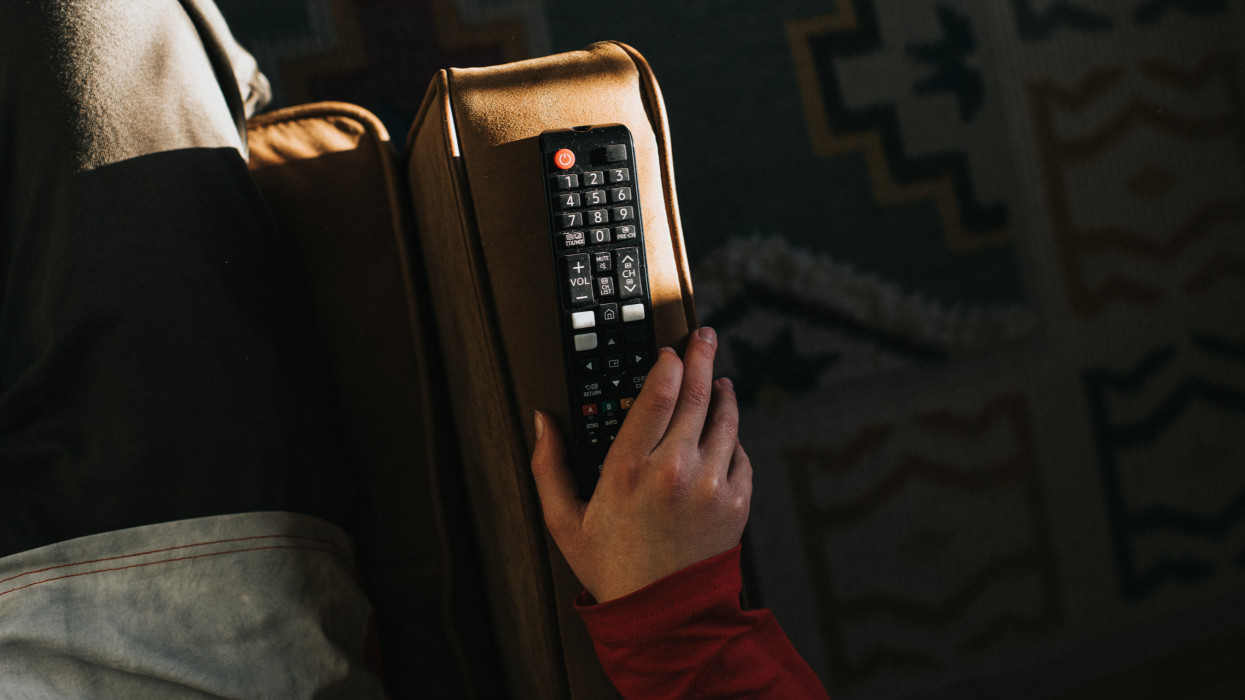 A black television remote control on the arm of a brown leather sofa. A hand grips the remote. Light shines in illuminating the various buttons. Space for copy.