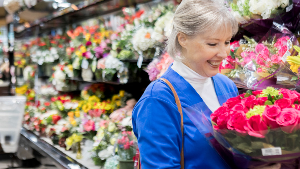 A smiling senior woman looks down at a bouquet of roses as she stands in the floral sections of a supermarket.