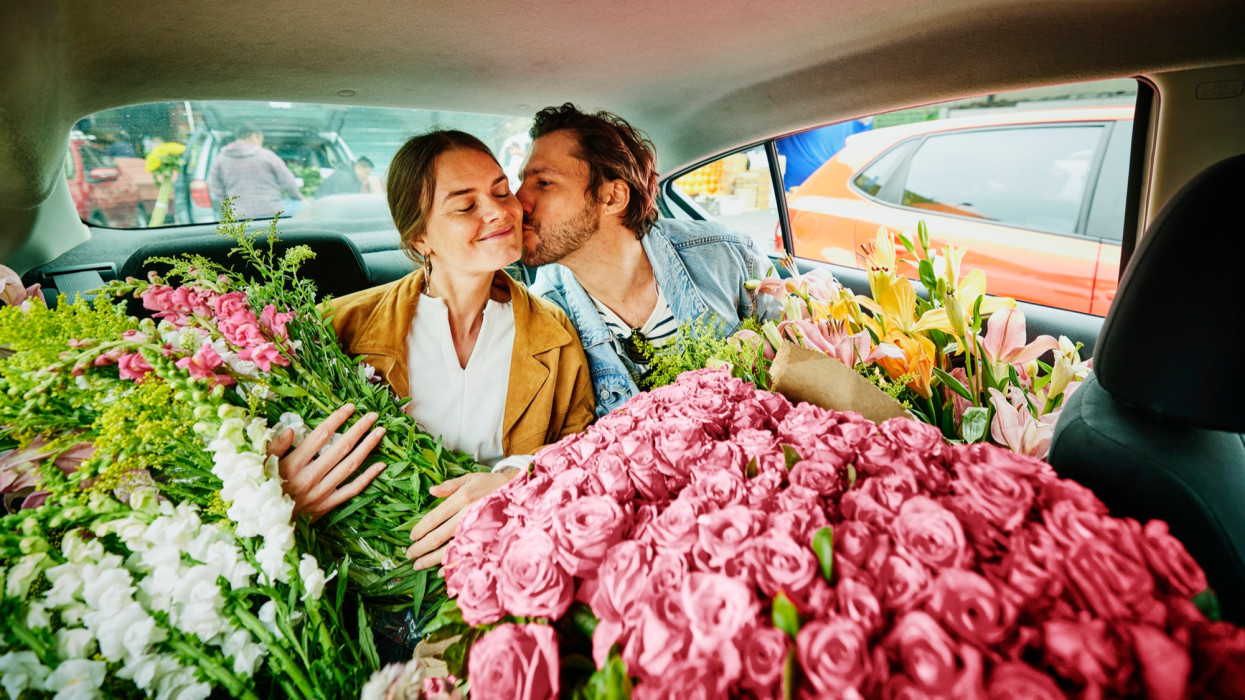 Medium shot of man kissing girlfriend on cheek in back of taxi filled with flowers after shopping at flower market during vacation
