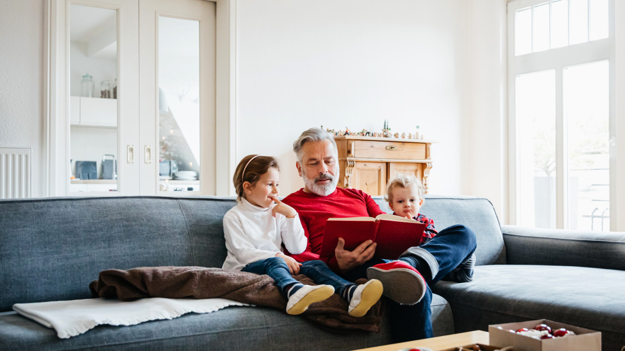 Two young grandchildren sitting on a large sofa and reading a festive book with their grandpa, getting ready for Christmas.