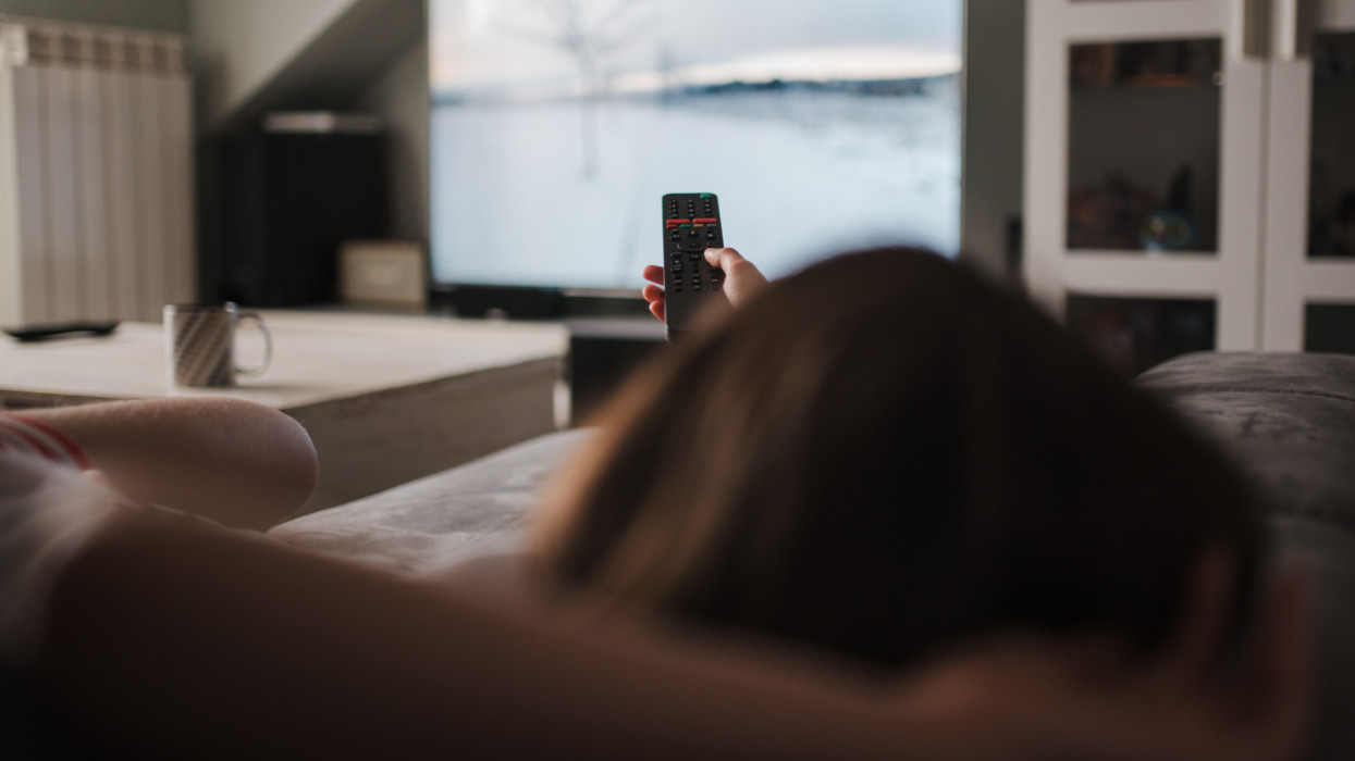View from behind of a young Caucasian girl lying on the sofa holding the TV remote control. She is relaxing on the sofa at home. Unrecognizable girl.