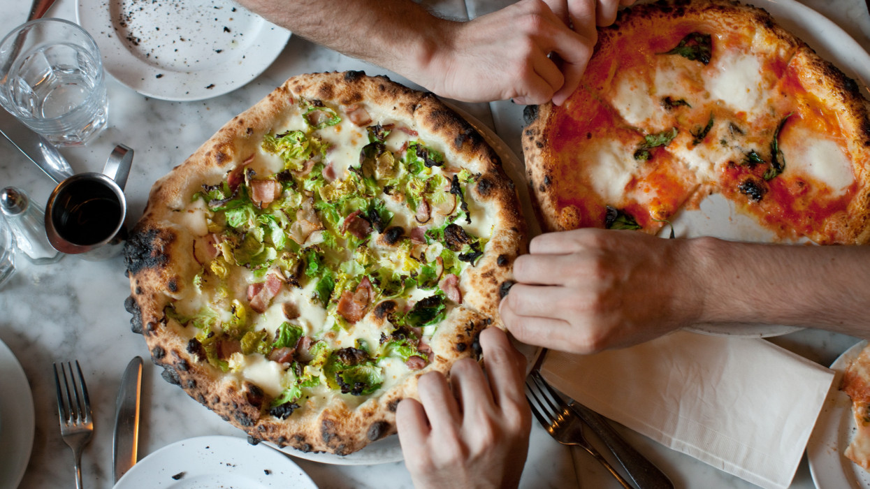Hands dig in to two pizzas (1 margherita, 1 with brussels sprouts and pancetta) on a marble table.