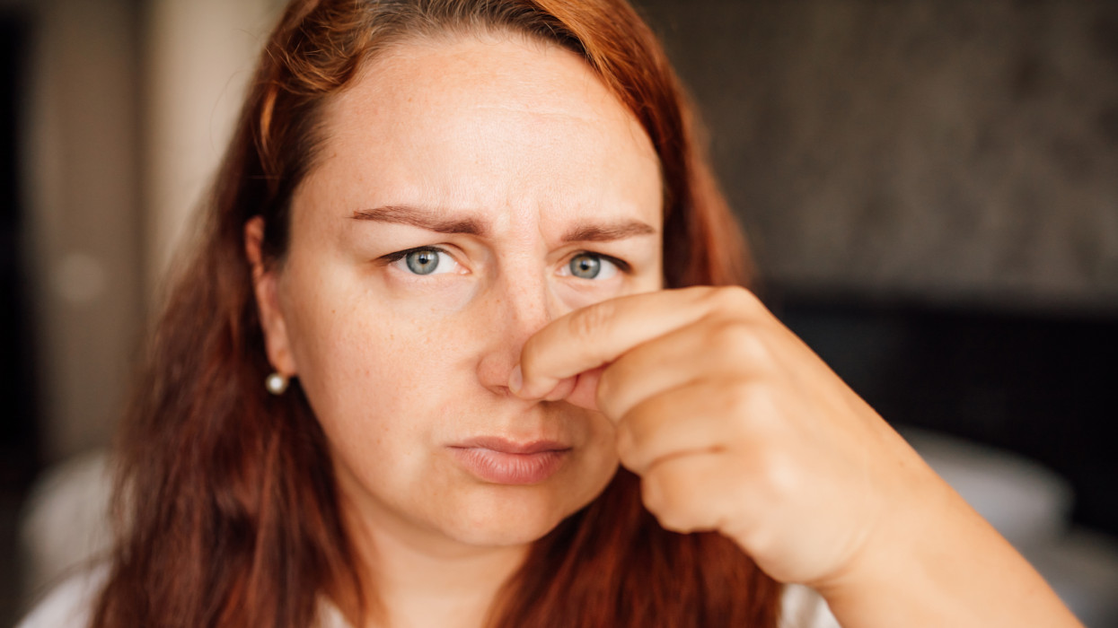 Photo of attractive mature redhead lady standing in bedroom with fingers gently pinching nose, displaying heightened smell sensitivity, as woman reacts to even faintest of scents, showcasing acute olfactory perception