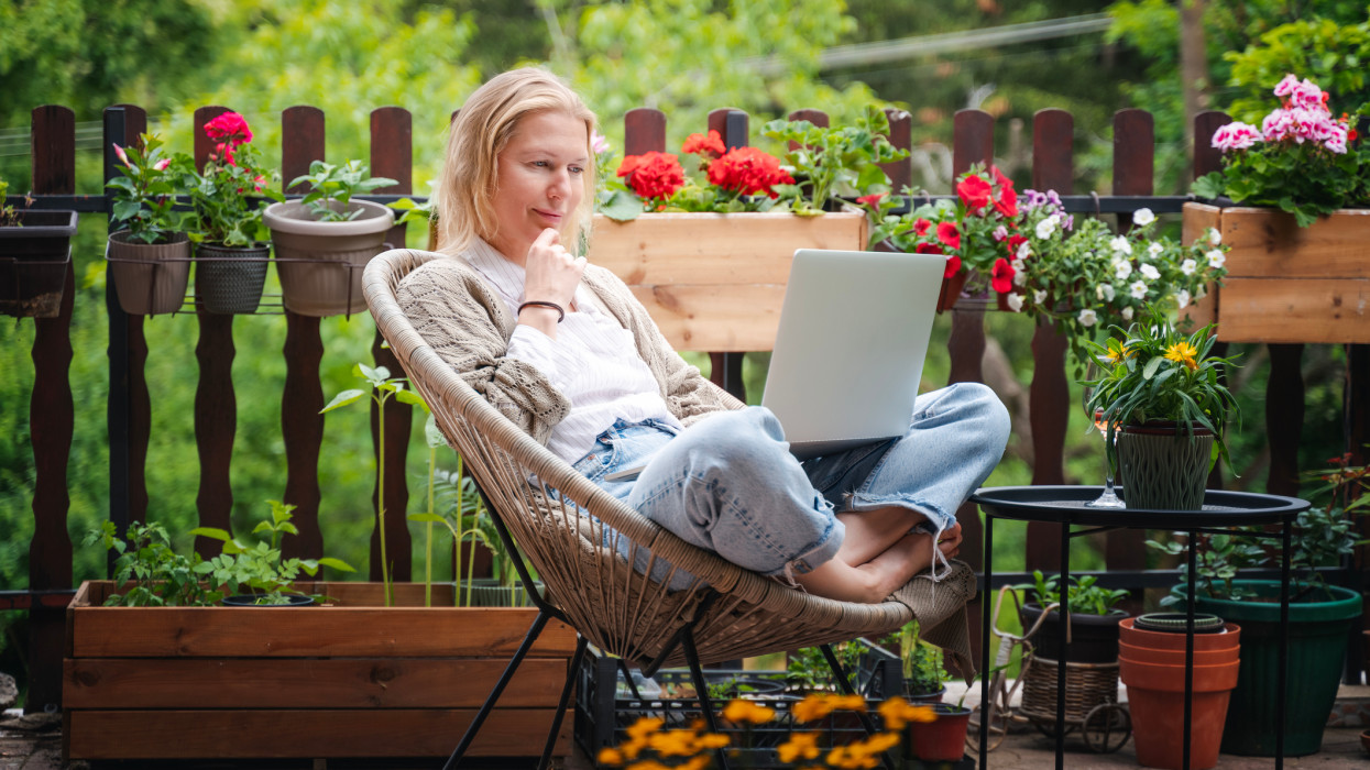 Beautiful blond middle-aged woman relaxing on the terrace of a country house in summer and using a laptop for online work balcony flower news