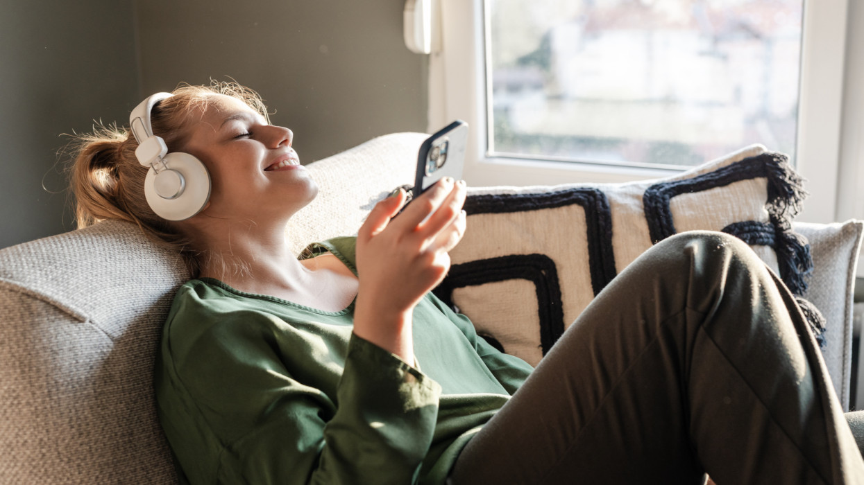 Young woman relaxing on a sofa and listening music on her phone via headphones