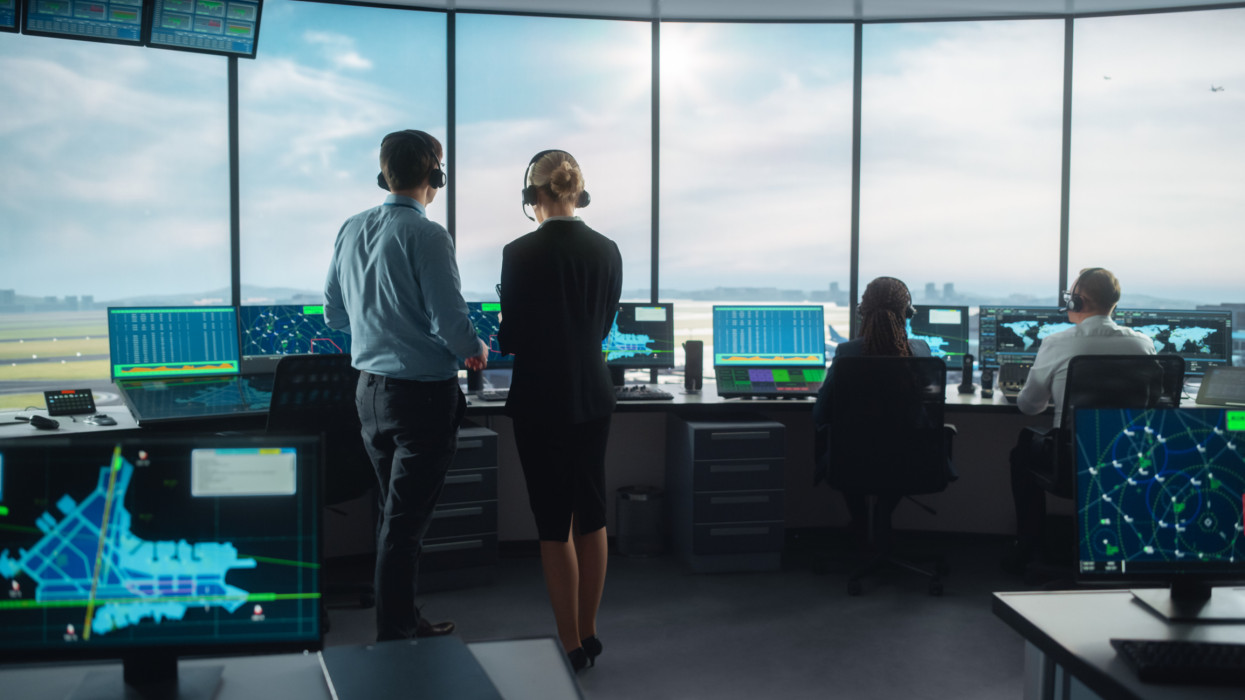 Female and Male Air Traffic Controllers with Headsets Talk in Airport Tower. Office Room is Full of Desktop Computer Displays with Navigation Screens, Airplane Departure and Arrival Data for the Team.