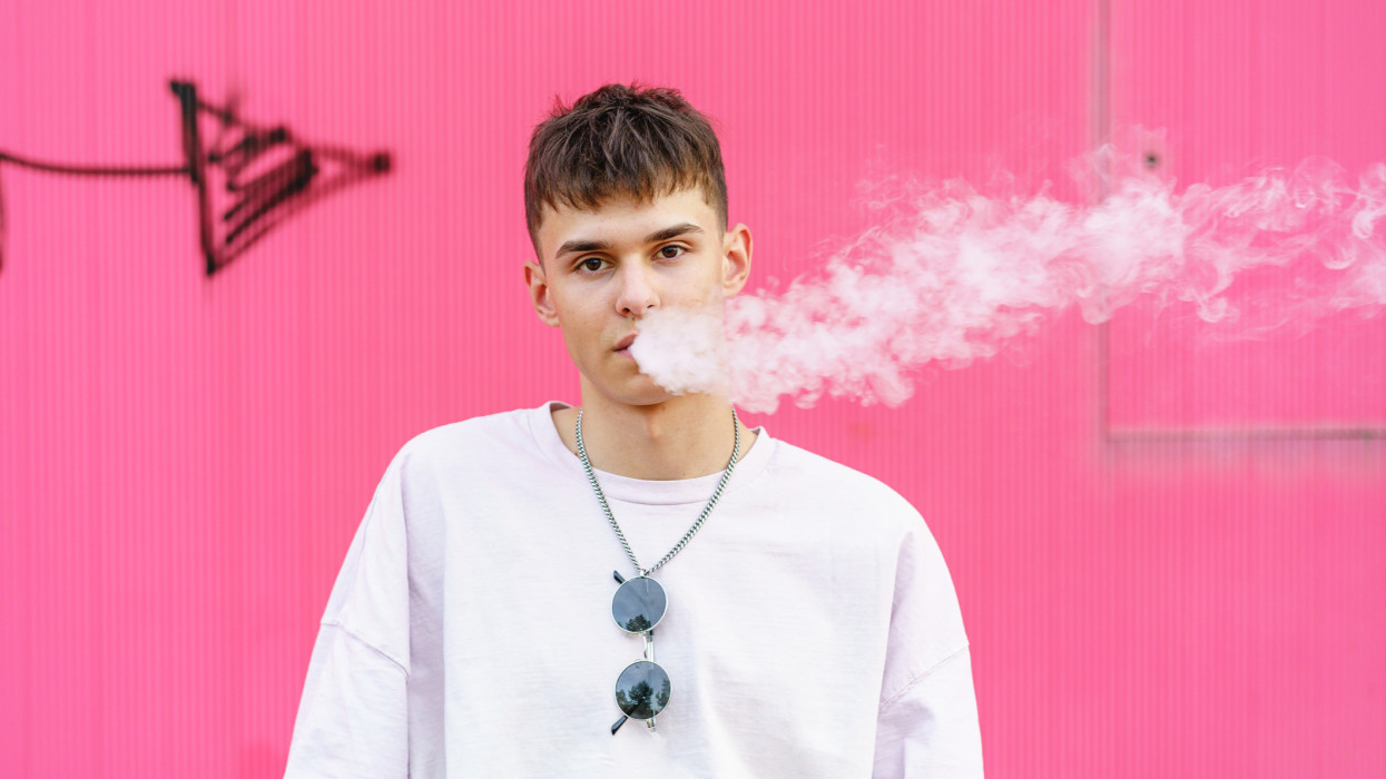 Horizontal cropped portrait of caucasian young man vaping white smoke isolated on pink urban background with graffiti. Diversity people lifestyles