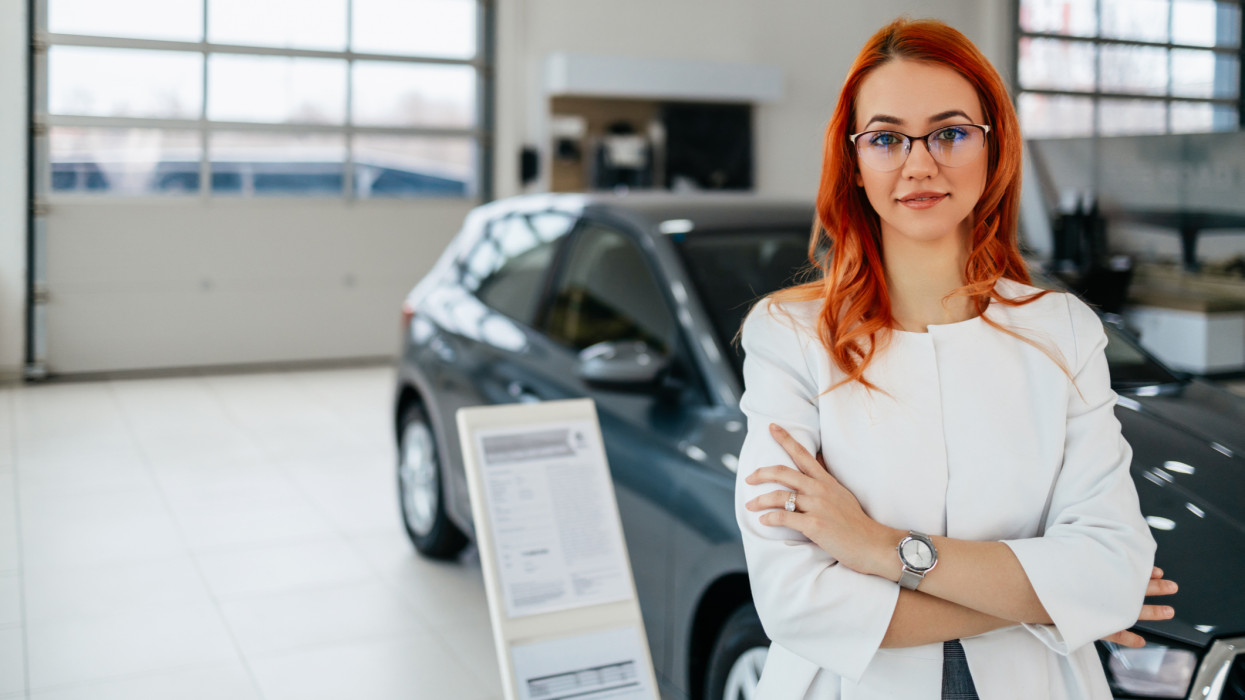 Positive young redhead saleswoman standing in car showroom and confidently looking at camera with arms crossed.