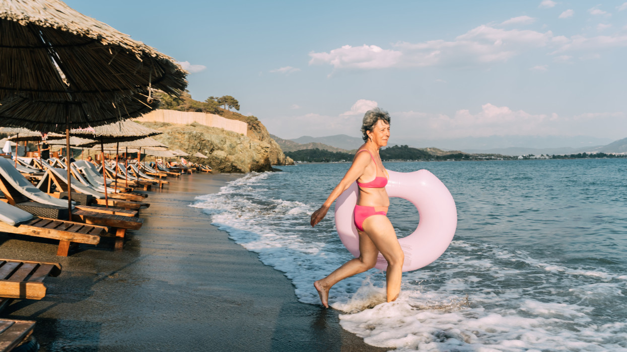Retirement-age woman wades into the sea with a pink inflatable circle donut.