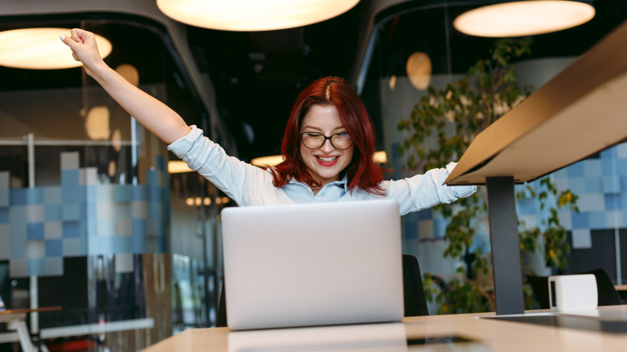 A beautiful young woman, who could be a design worker, trainee, or businesswoman, exudes joy and celebration in an open, modern office work area. With her arms extended outwards, she signifies a moment of triumph, possibly after a successful business deal.Business deal, beautiful, young woman, design worker, trainee, businesswoman