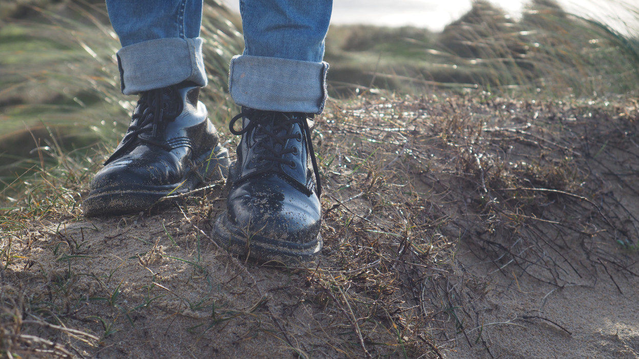 Dr Martens boots with jean pant, on the sand and grass of a beach. Melancolic vibe, sea in the background