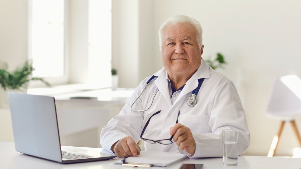 Portrait of happy, friendly, trustworthy, experienced senior doctor looking at camera sitting at desk with laptop computer in modern hospital office. Healthcare, professional medical help, expertise