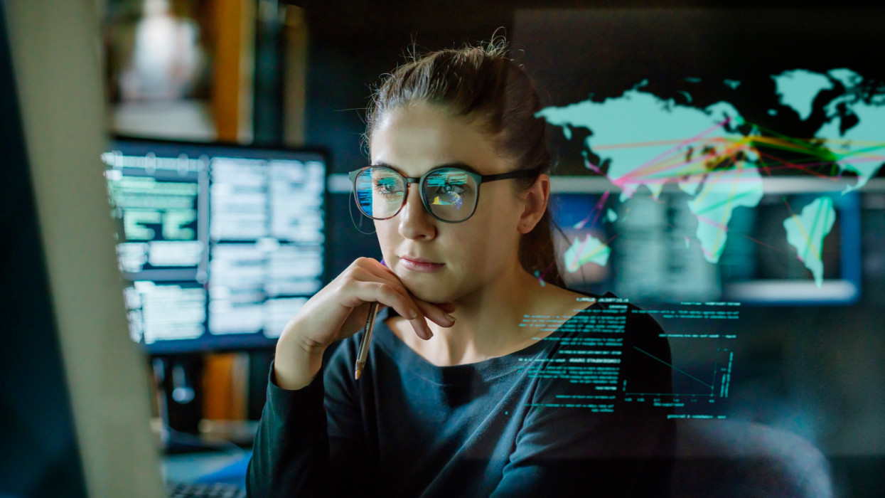 Stock image of a young woman, wearing glasses, surrounded by computer monitors in a dark office. In front of her there is a see-through displaying showing a map of the world with some data.