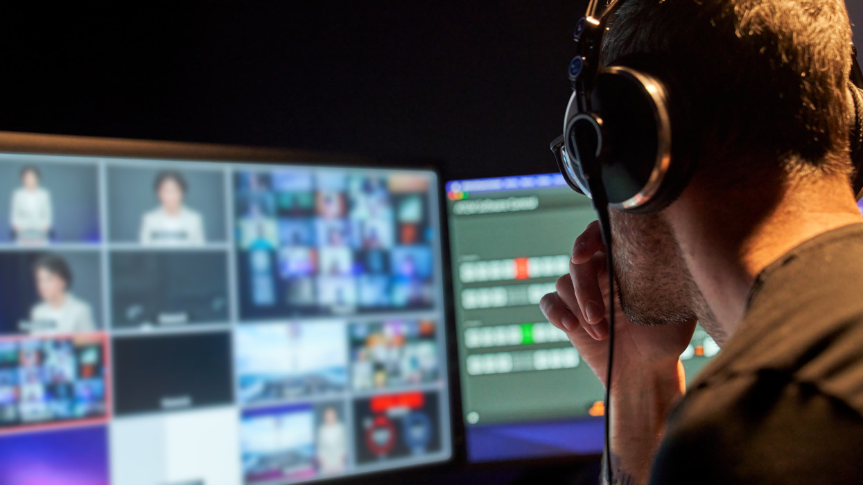 Rear view of television producer working during filming, live streaming or live broadcast. He is concentrated looking at the computer screen controlling the cameras using software.