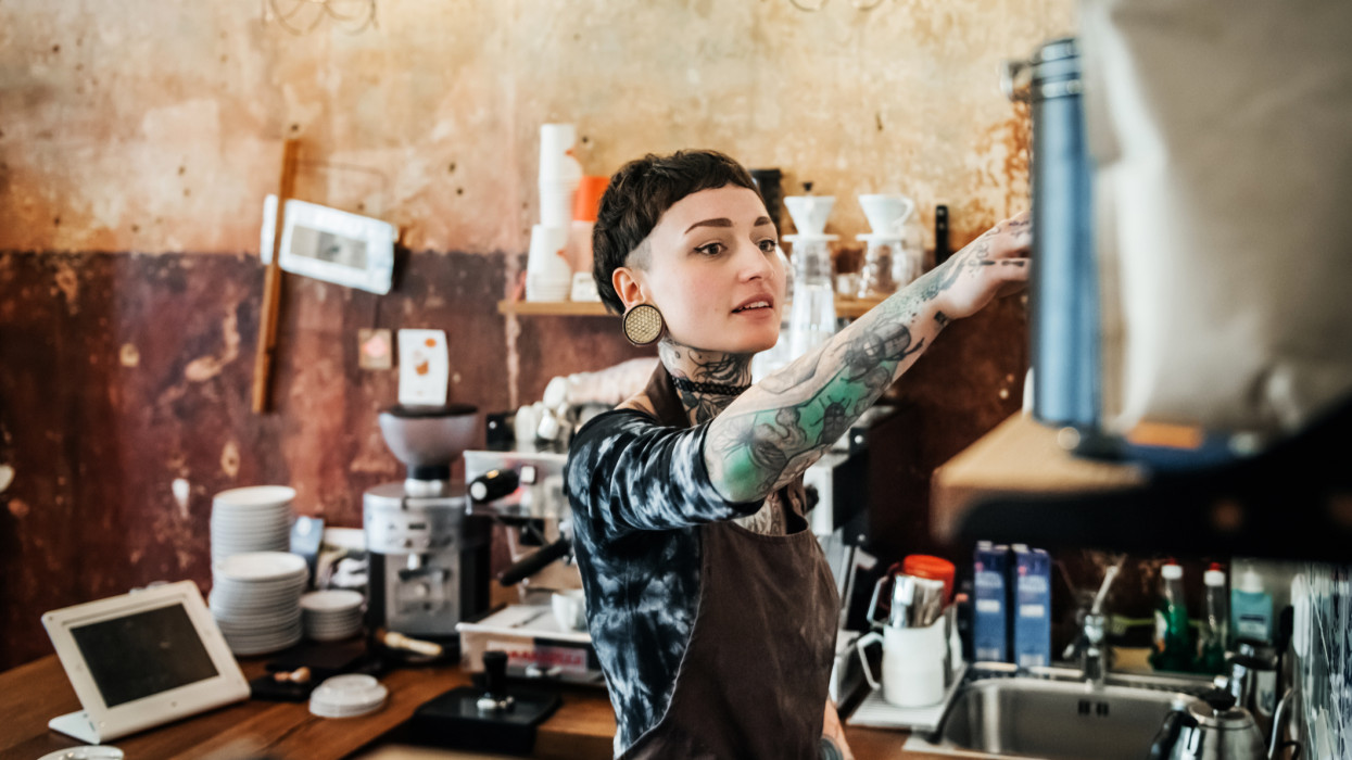 A young tattooed woman wearing an apron is working as a waitress in a bright cafÃ©. She is happily smiling while grabbing something.