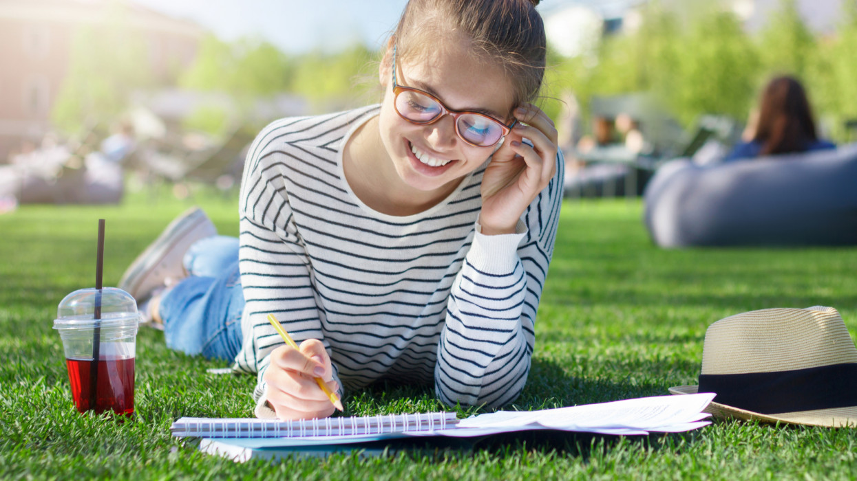 Closeup of good-looking student girl on grass in park, her head bent towards notebooks where she writes answers, smiling, drinking berry juice and feeling happy with getting ready for studies