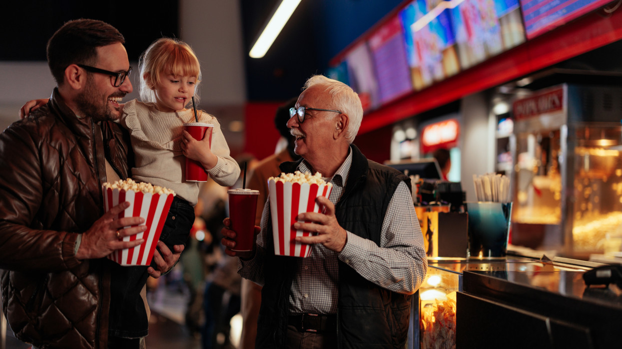 A cheerful grandfather is in the cinema with his son and granddaughter, holding popcorn and beverages in their hands on their way to see a movie.
