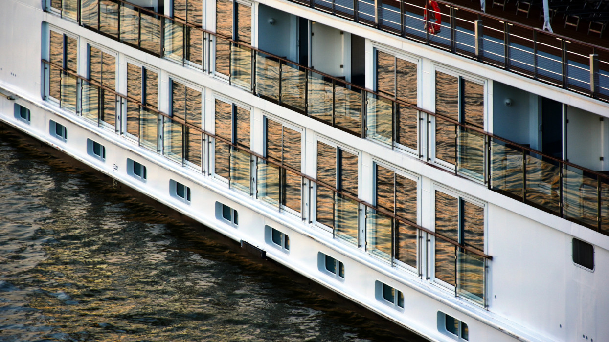large white multi level hotel boat on the Danube river at sunset. warm brown green tones and reflections on the water. top deck and glass balustrade. hospitality, accommodation and sightseeing. leisure, travel and tourism concept