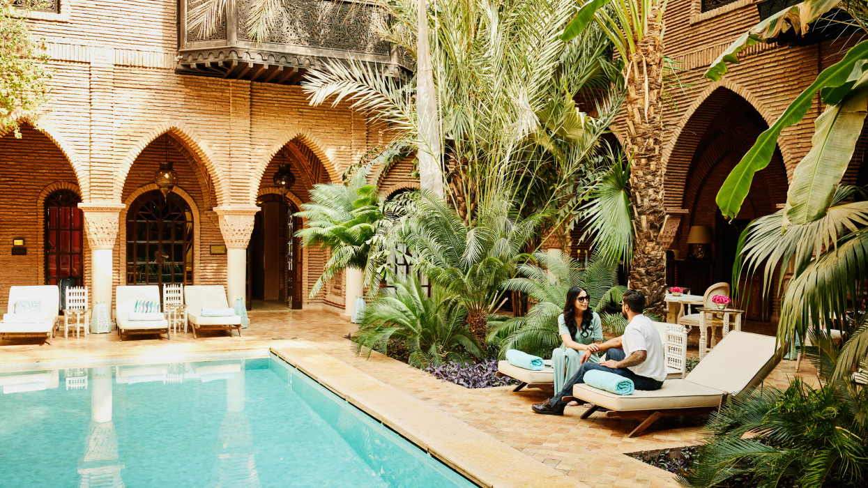 Wide shot of smiling couple sitting by pool in courtyard of luxury hotel while on vacation