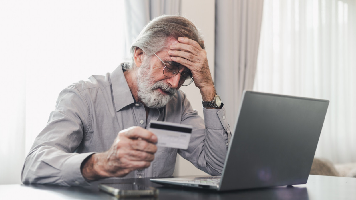 Zaklatott idős férfi, aki hitelkártyát tart a laptop előtt, gondjai vannak a pénzügyi biztonsági adatokkal vagy az online fizetés biztonságával. Banki ügyfél hitelkártya miatt aggódik, pénzügyi csalás áldozata lett