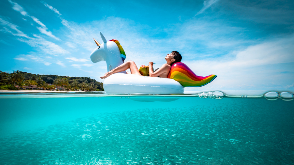 Girl holding a coconut enjoying the sun lying on a floating unicorn with fish swimming underwater