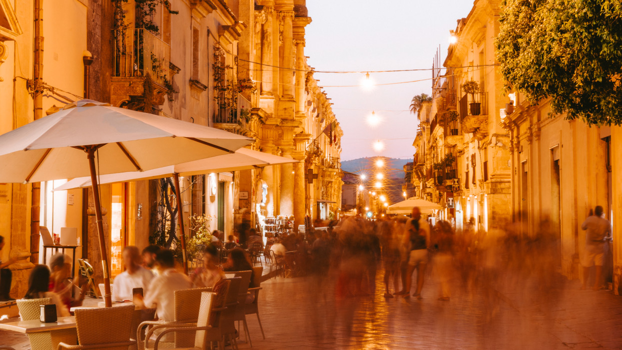 Italy, Sicily, Noto, people dining and socialising outside at dusk in busy street