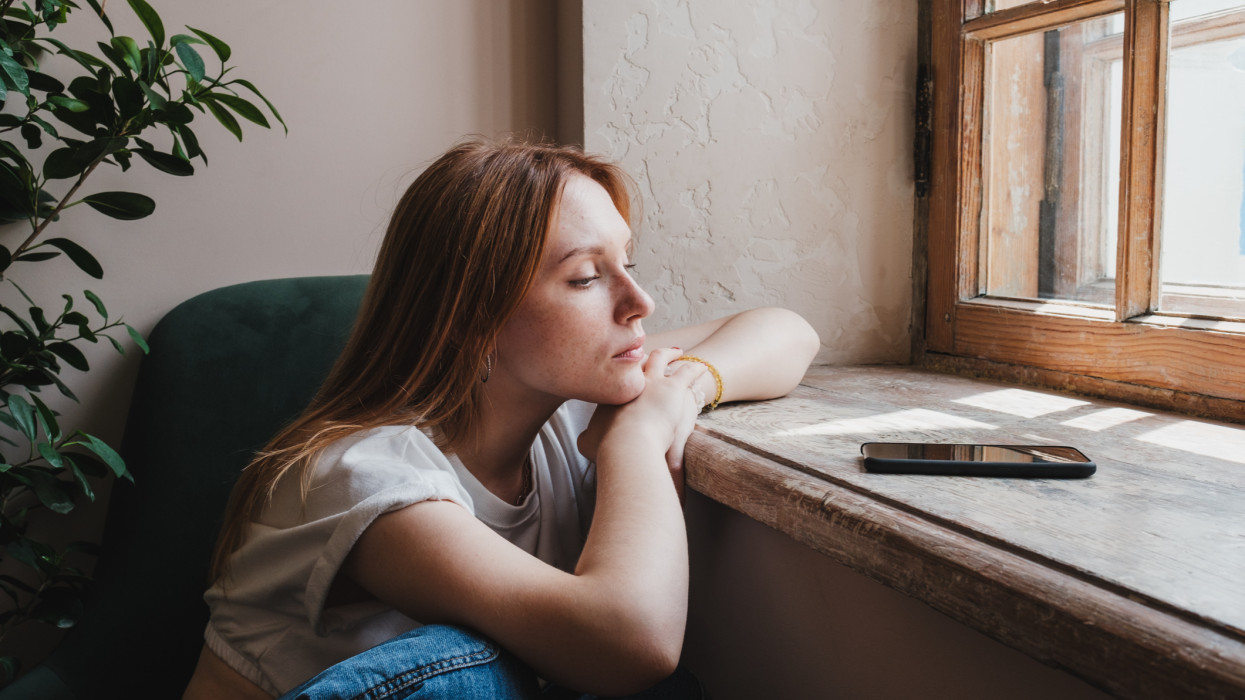 Upset redhead teen girl sitting by window looking at phone waiting call from boyfriend, feeling sad and depressed teenager looking at smartphone wait for message. Social Media depression in teens