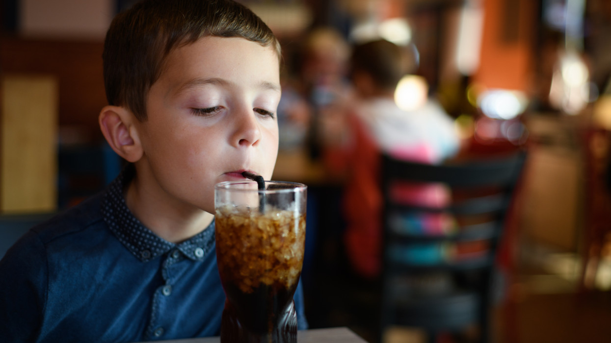 Image of a six year old boy drinking a sugary drink (cola) through a straw sitting at a table in a restaurant.