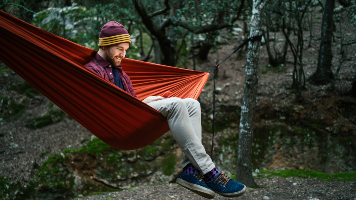 Young male uses his smartphone resting on a hammock in the woods