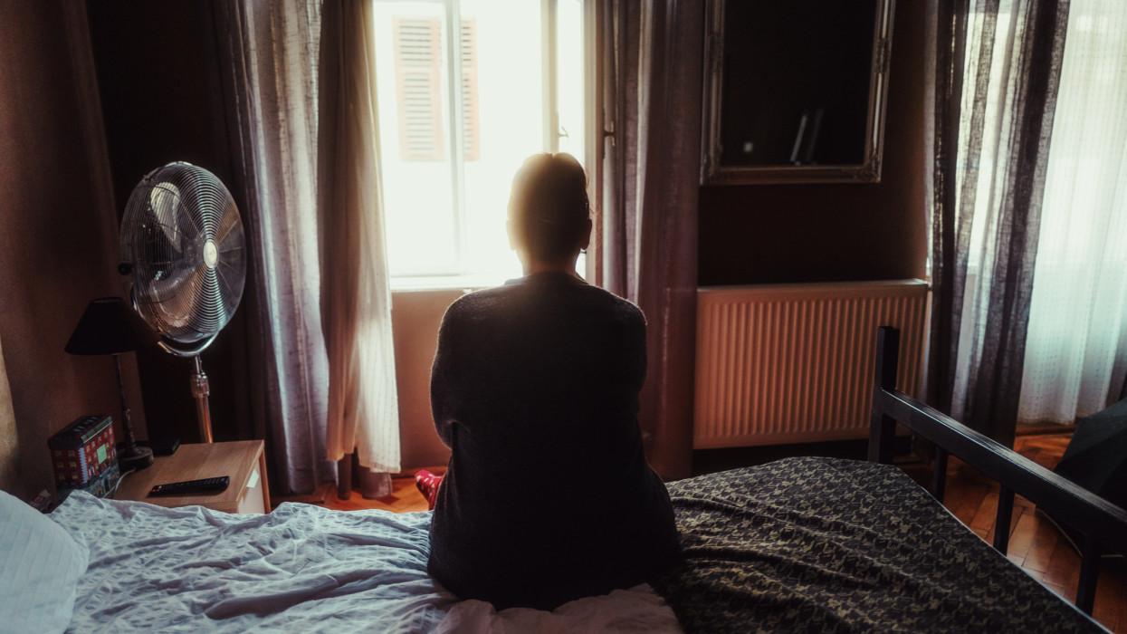Color image depicting the rear view of a mid adult woman sitting on the edge of her hotel room bed.