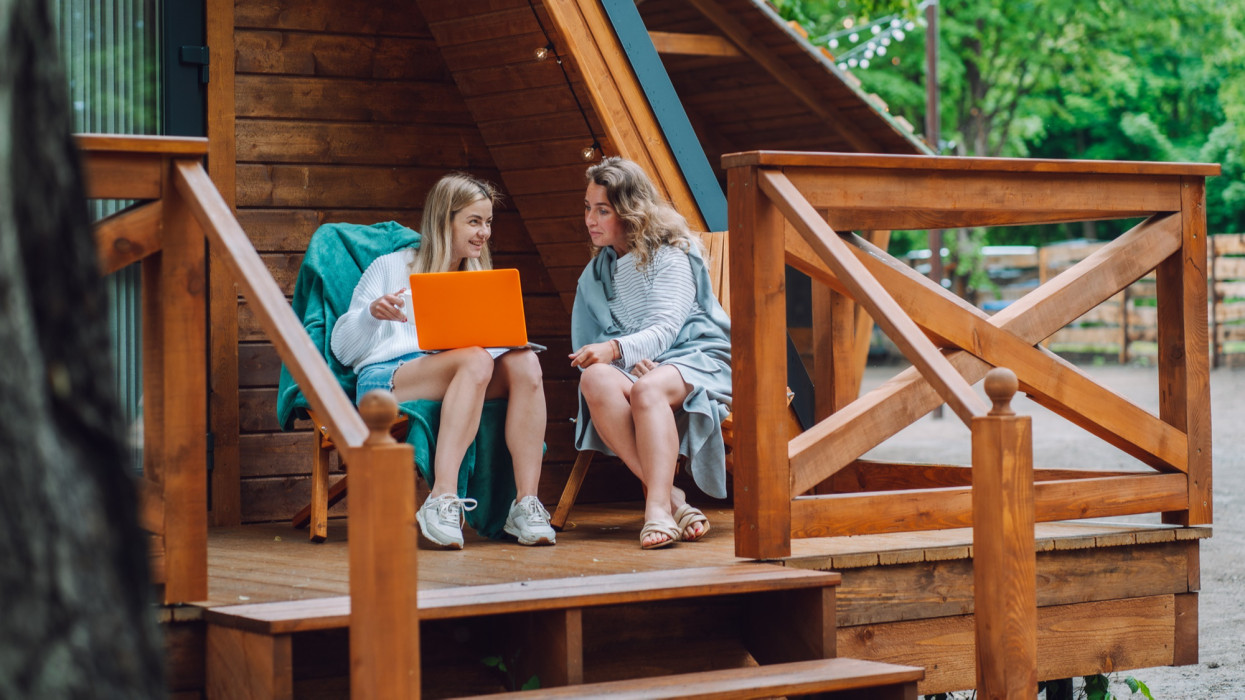 Two women using orange laptop and relaxing on wooden house patio outdoors. Glamping retreat and getaway for digital nomads and freelancers