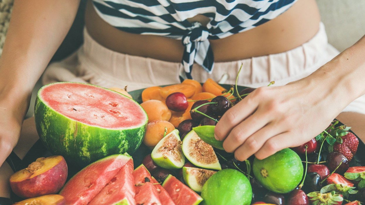 Summer healthy raw vegan clean eating breakfast in bed concept. Young girl wearing striped home shirt sitting and taking fruit from tray full of fresh seasonal fruit, square crop