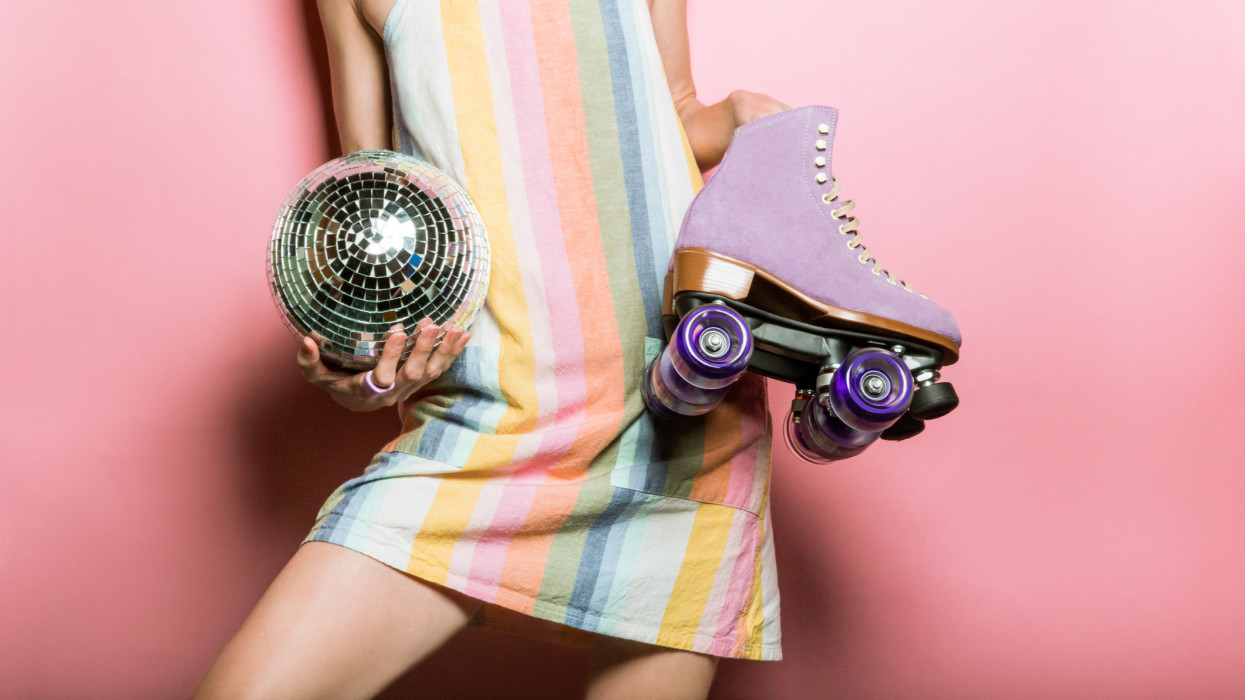 Young woman holding trendy rollerskates and disco ball. Retro 1970s woman is wearing a pastel striped dress and is holding purple roller skates against pink background with copy space. Conceptual image for fashion trends, pandemic hobbies, and 70s parties.