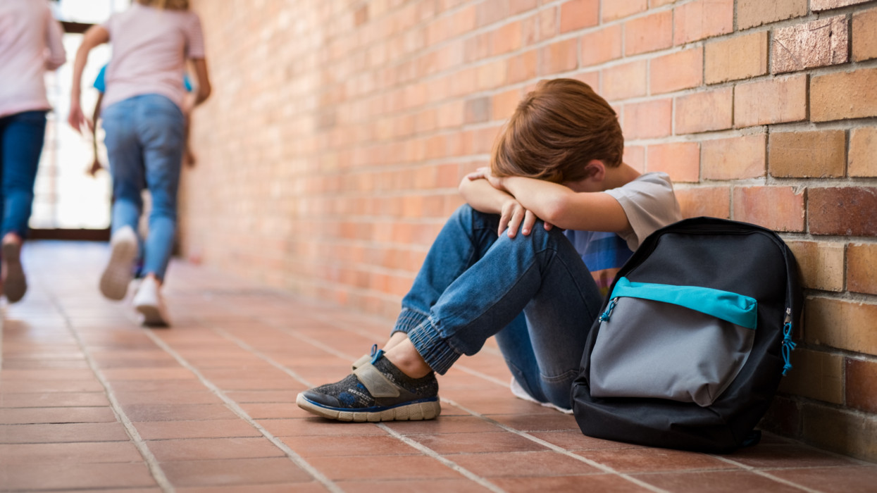 Little boy sitting alone on floor after suffering an act of bullying while children run in the background. Sad young schoolboy sitting on corridor with hands on knees and head between his legs.