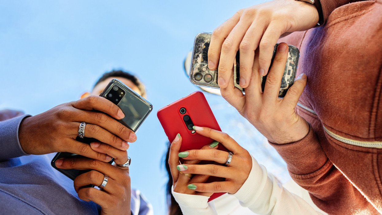 Low angle view of three young people using mobile phones outdoors. Addicted gen z friends holding smartphone outside. Blue sky. Copy space for text.