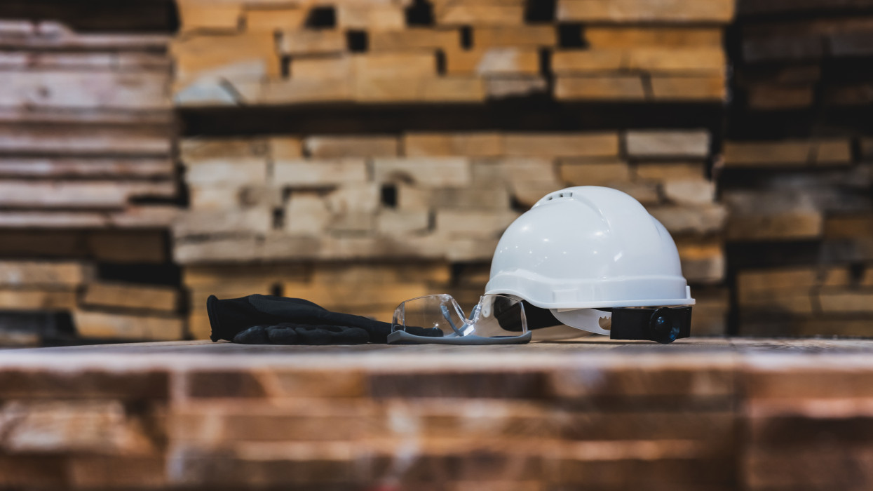 White hard hat, safety glasses, and work gloves sit on a stack of lumber, representing workplace safety and preparedness in industrial settings