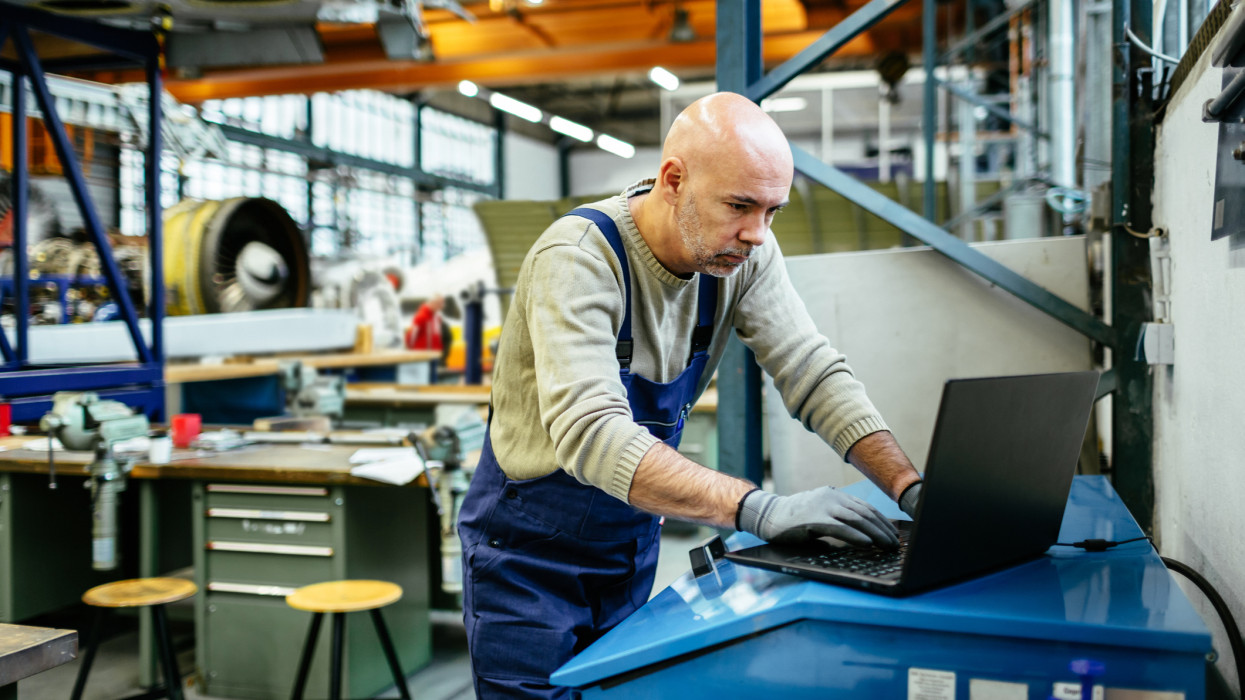 Egy érett repülőgép-szerelő dolgozik egy laptopon egy hatalmas repülőgép-hangárban. Munkaruhát visel, mint egy overallt, miközben a háttérben gépek és motorok láthatók.