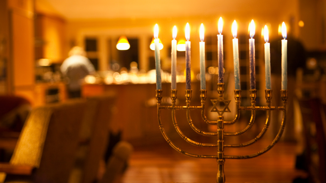Lit candles in a menorah with a women working in the kitchen in the background, commonly used during the Jewish celebration of Hanukkah.