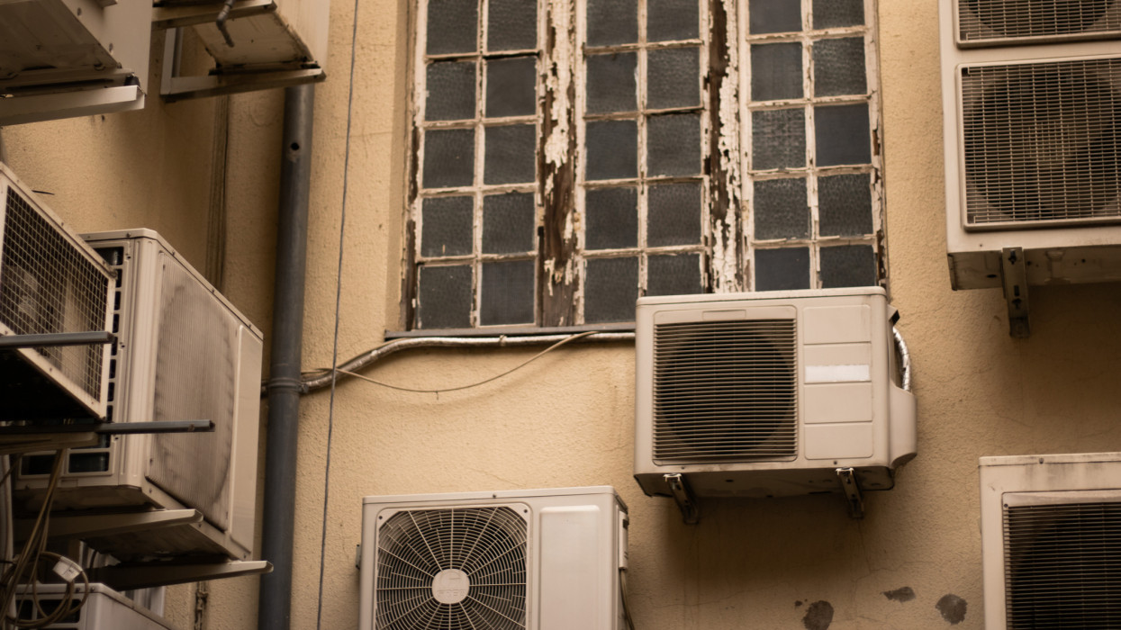 Egy elöregedett, hámló festékkel ellátott ablak, amelyet több, bézs színű falra szerelt légkondicionáló egység vesz körül. A jelenet egy városi környezetet örökít meg a kopás és a használat látható jeleivel.